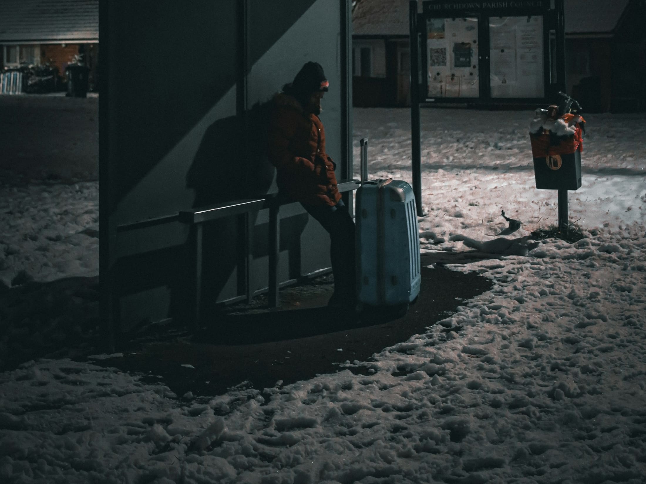 A person in a red parka waits alone at a snow-covered bus shelter at night, a blue suitcase beside them.